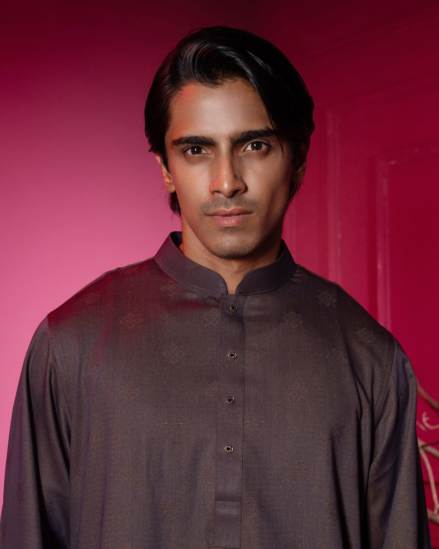 Close portrait of a man in a charcoal jacquard VIOR panjabi, lit against the red room wall